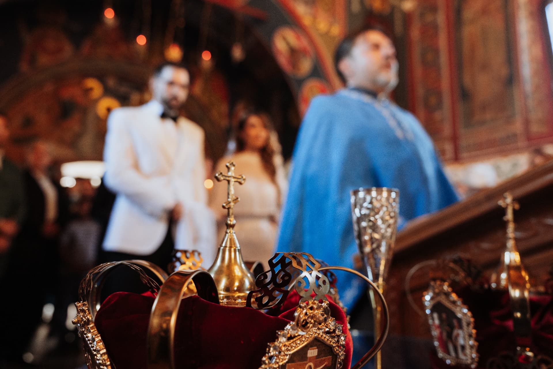 Golden wedding crowns on red velvet in the foreground, with the bride, groom and priest during the Orthodox church ceremony