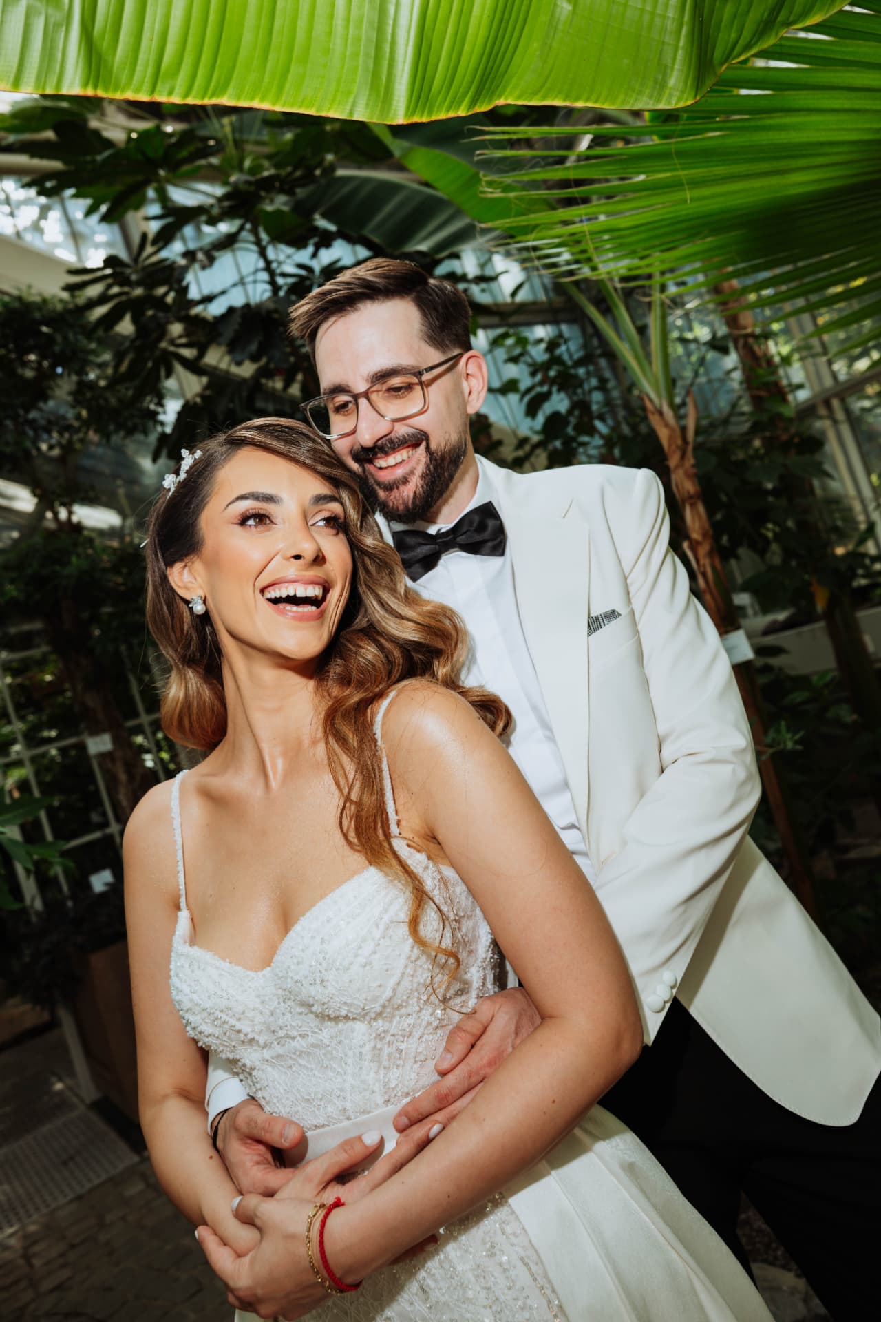 Bride laughing with her head turned away while the groom holds her, photographed under a large banana leaf in a botanical garden.