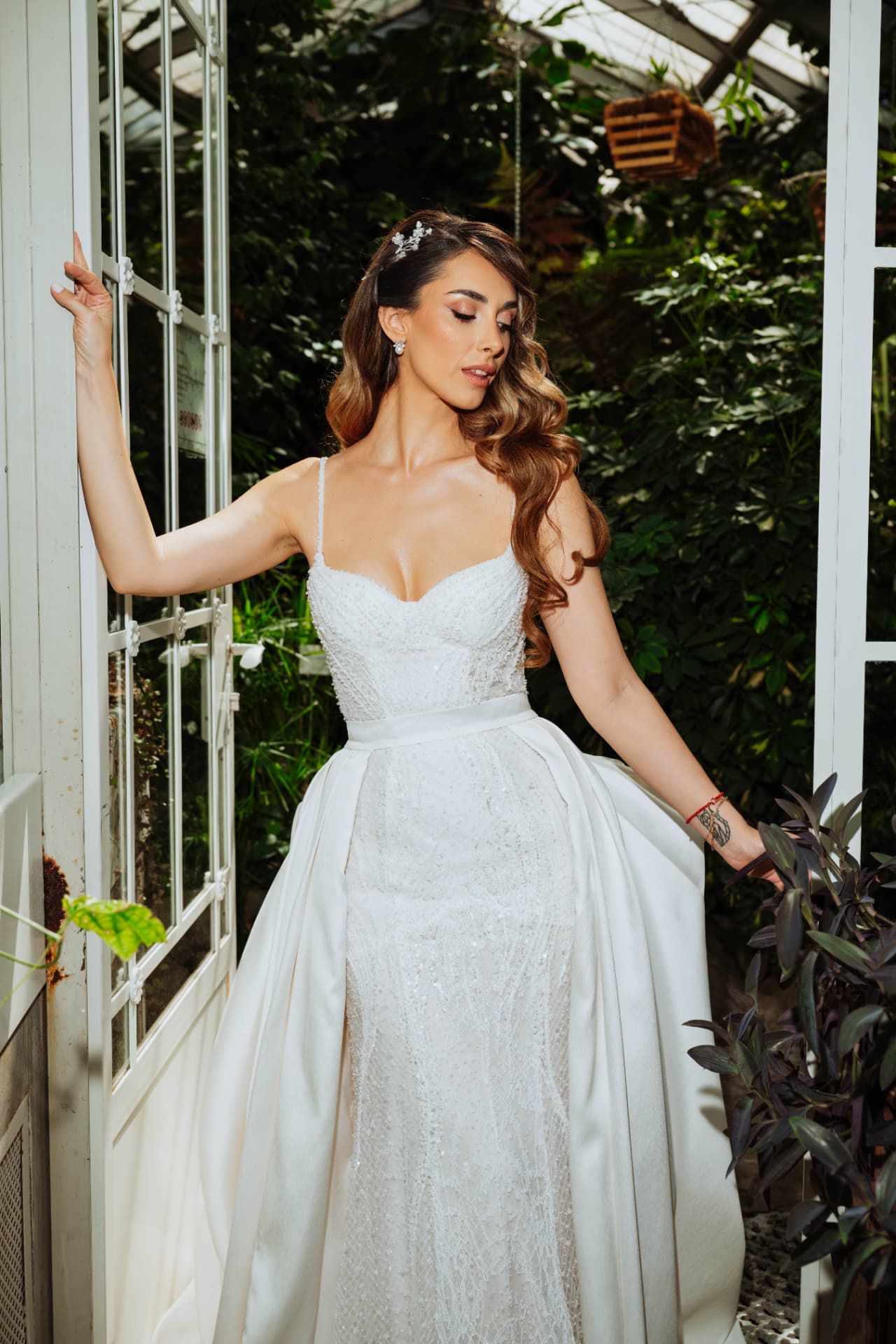 Bride in a beaded gown with an overskirt holding open a white greenhouse door, surrounded by dense green foliage.