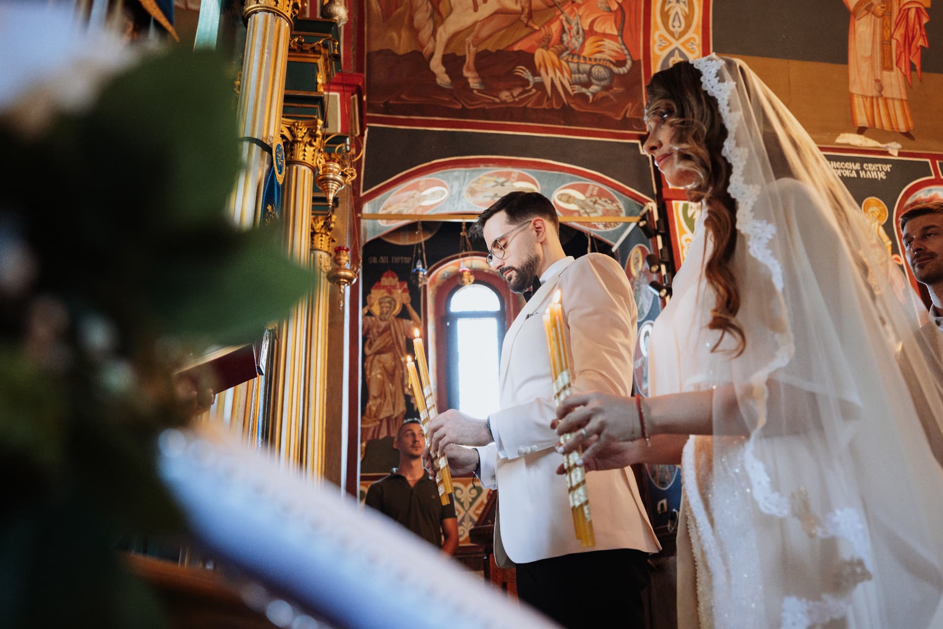 Jelena and Vladimir holding lit candles during their Orthodox wedding ceremony in a frescoed church in Serbia