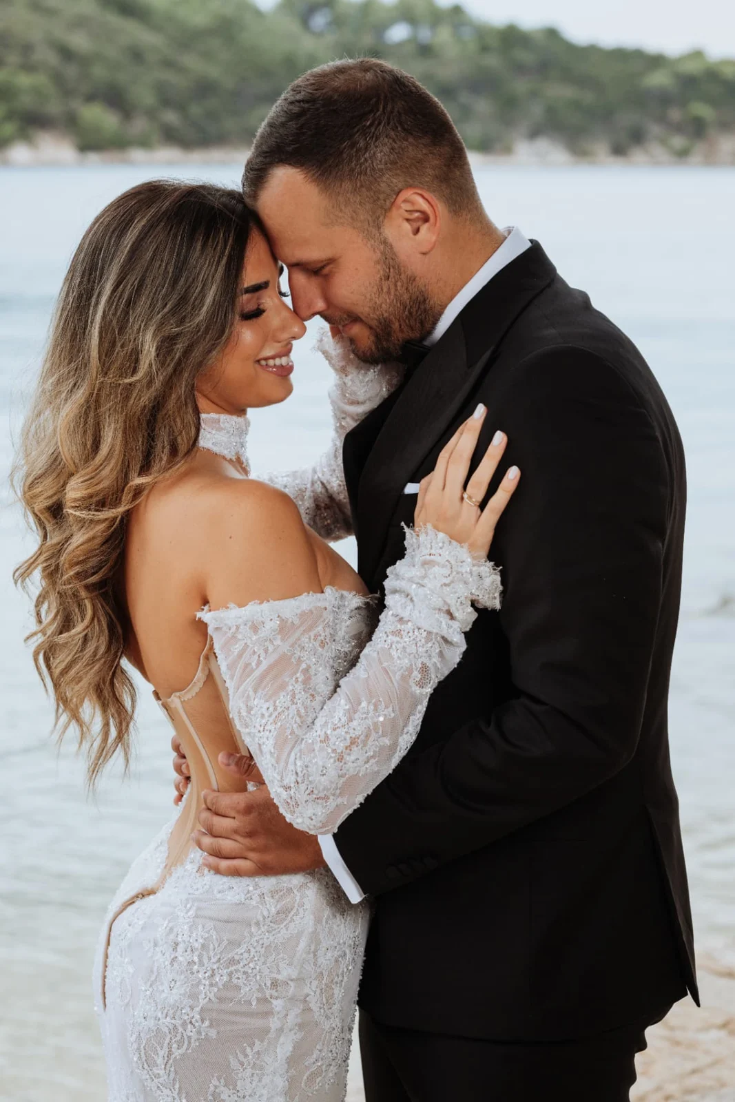 Bride and groom standing forehead to forehead by the sea, the bride smiling in a long-sleeved lace gown.