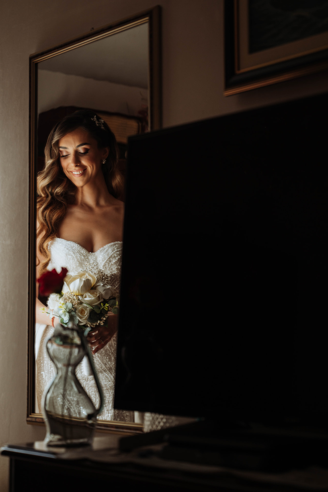 Bride reflected in a gilded mirror in a dim room, holding a bouquet of white roses and a single red flower.