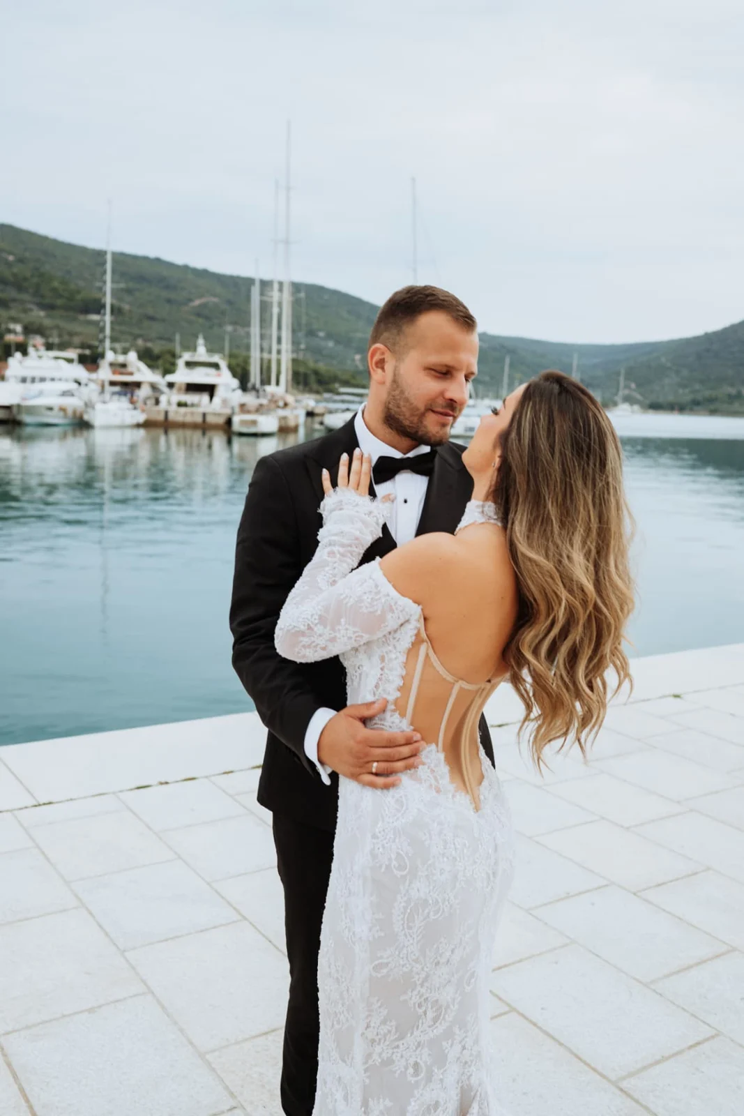 Bride with long wavy hair looking up at the groom in a black tuxedo on a stone waterfront, sailing boats in the harbour behind them.