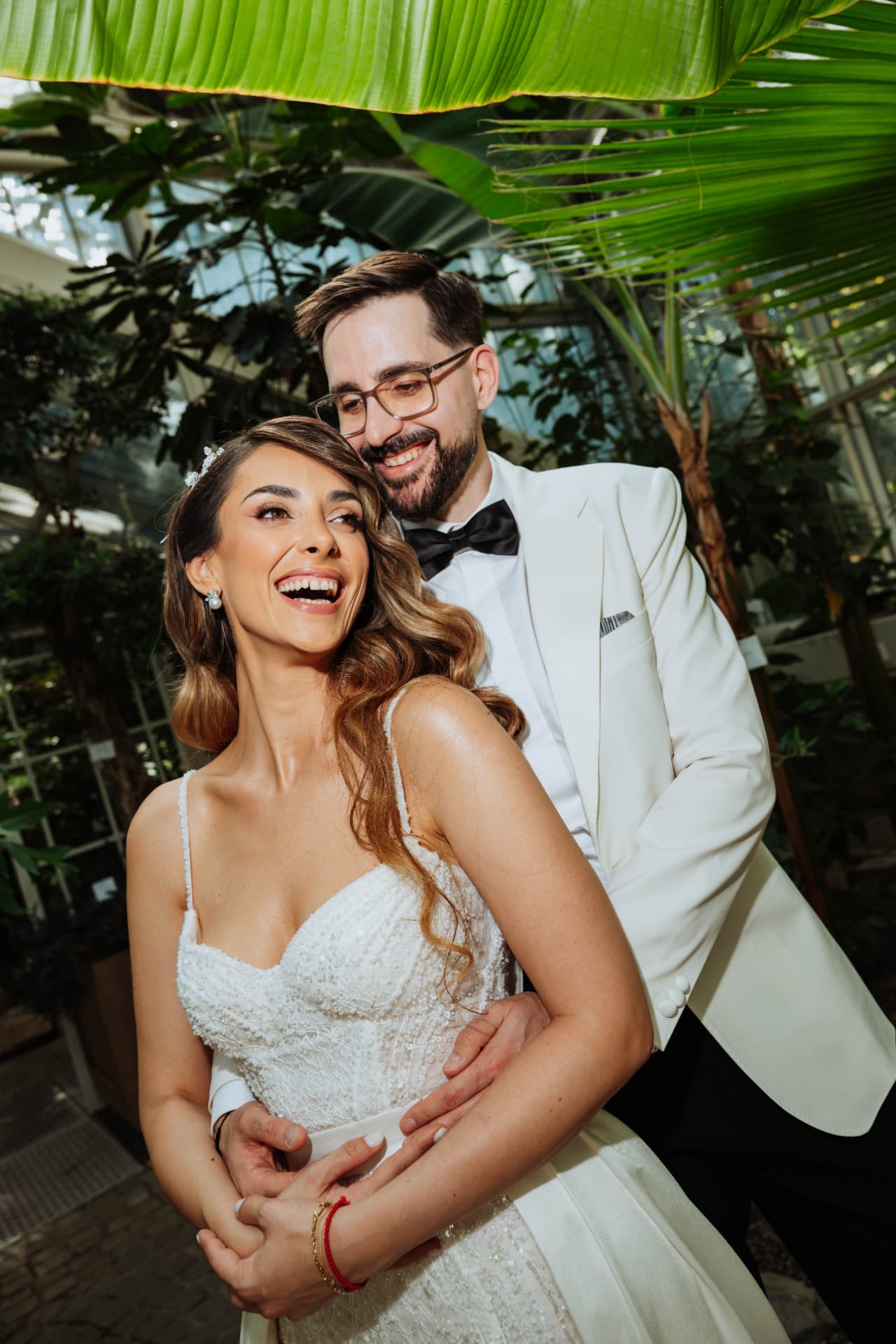 Bride laughing openly as the groom leans in behind her, framed by palm fronds inside a botanical glasshouse.